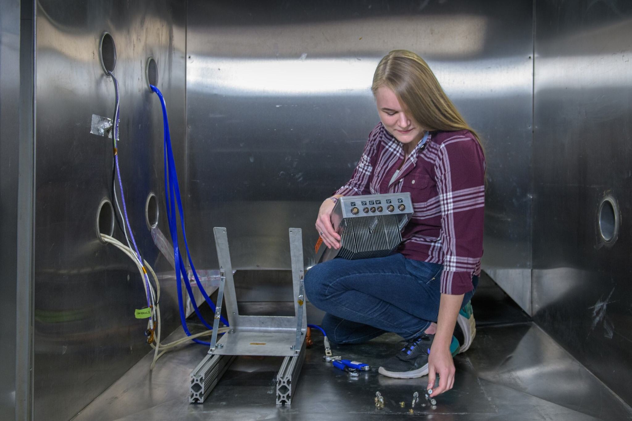 A woman wearing a flannel shirt and jeans kneels on the ground while holding a large metal device. Next to her is a metal apparatus with blue and white cables feeding through holes in the wall.