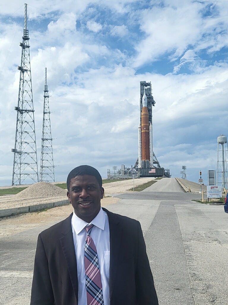 A man stands outside in front of a rocket. He is wearing a white button down