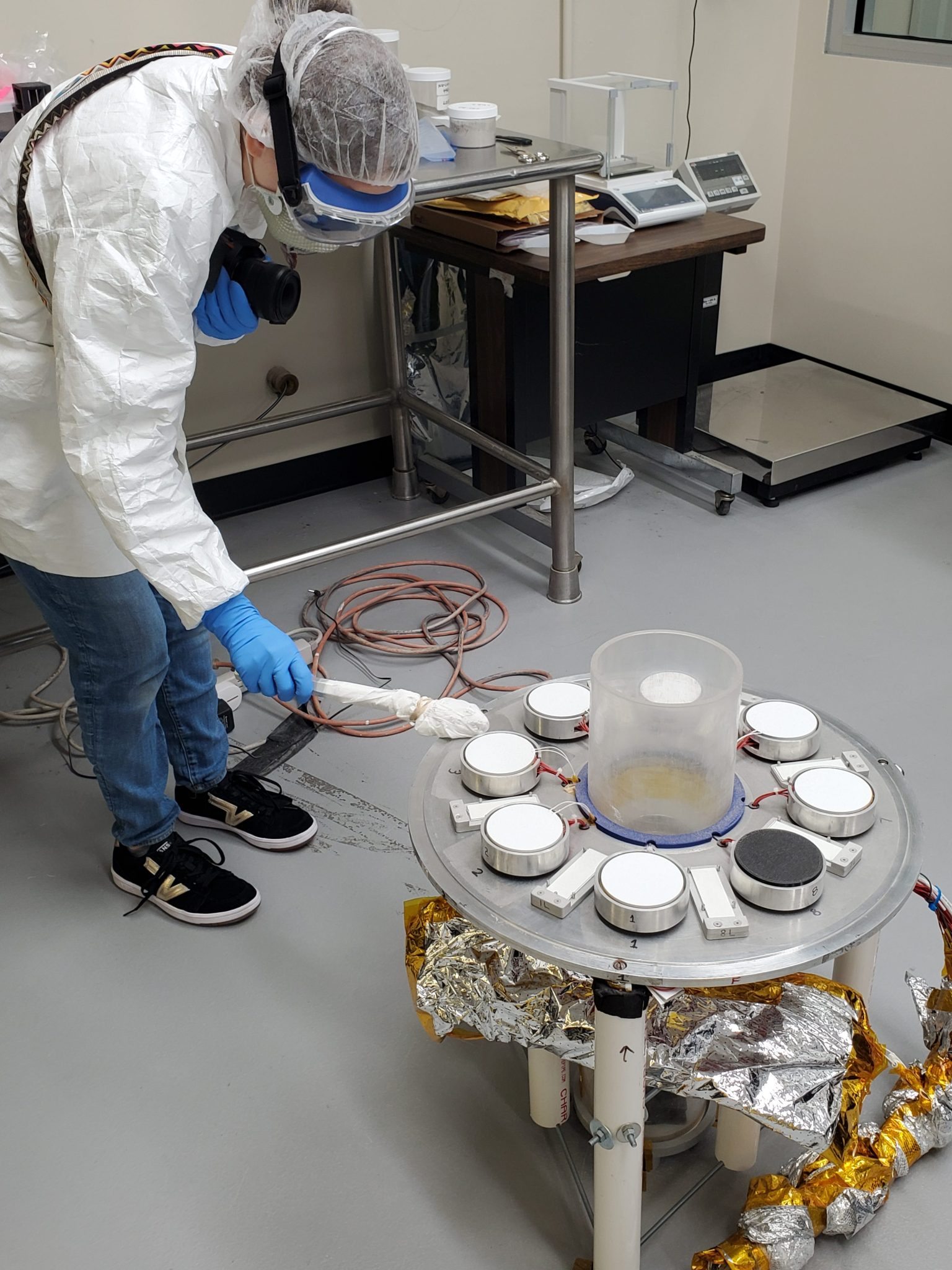 A NASA employee wearing a lab coat, hair net, and safety goggles conducts an experiment with lunar simulant on a round tray.