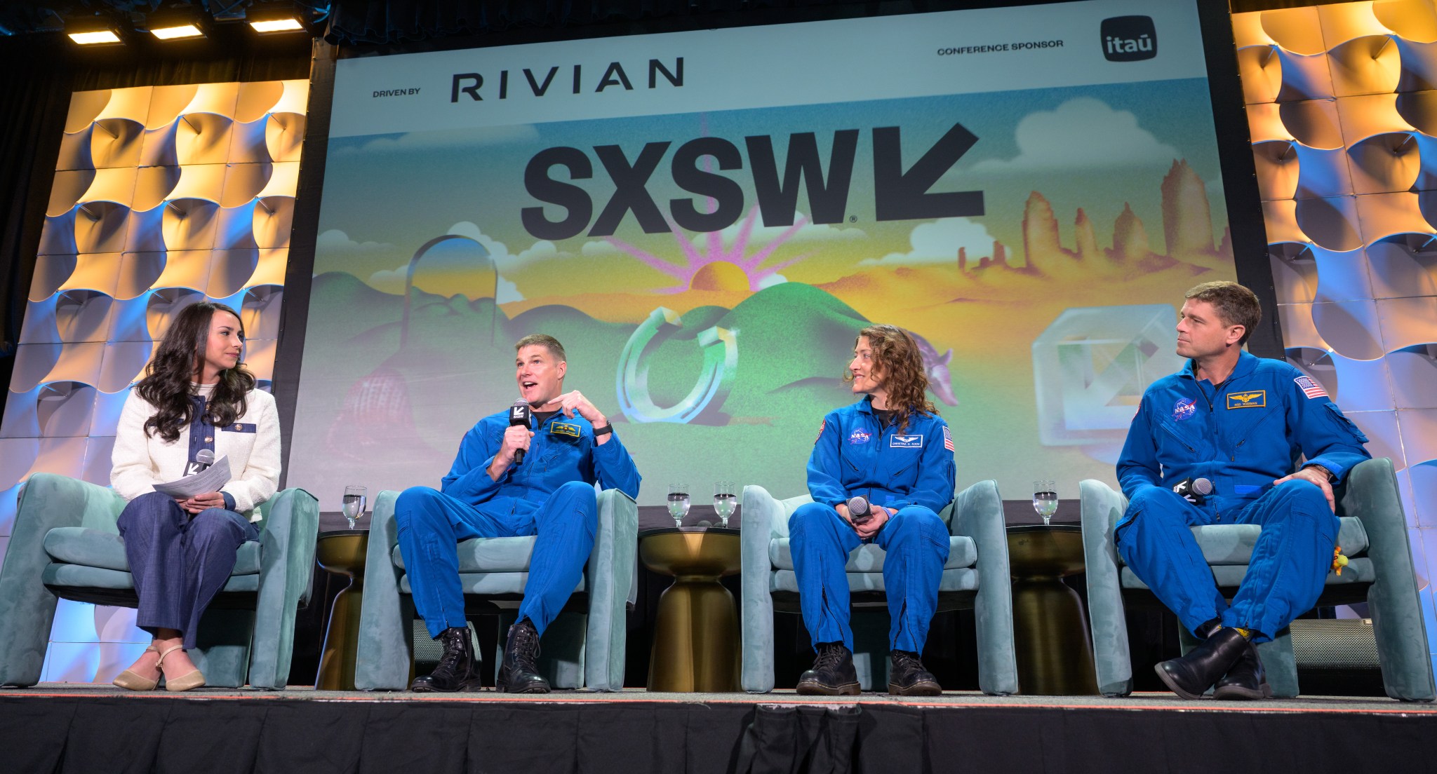 Two women and two men sit in teal velvet chairs on a stage. The SXSW logo is behind them on a screen. Three of the people on stage are wearing blue flight suits; they are astronauts.