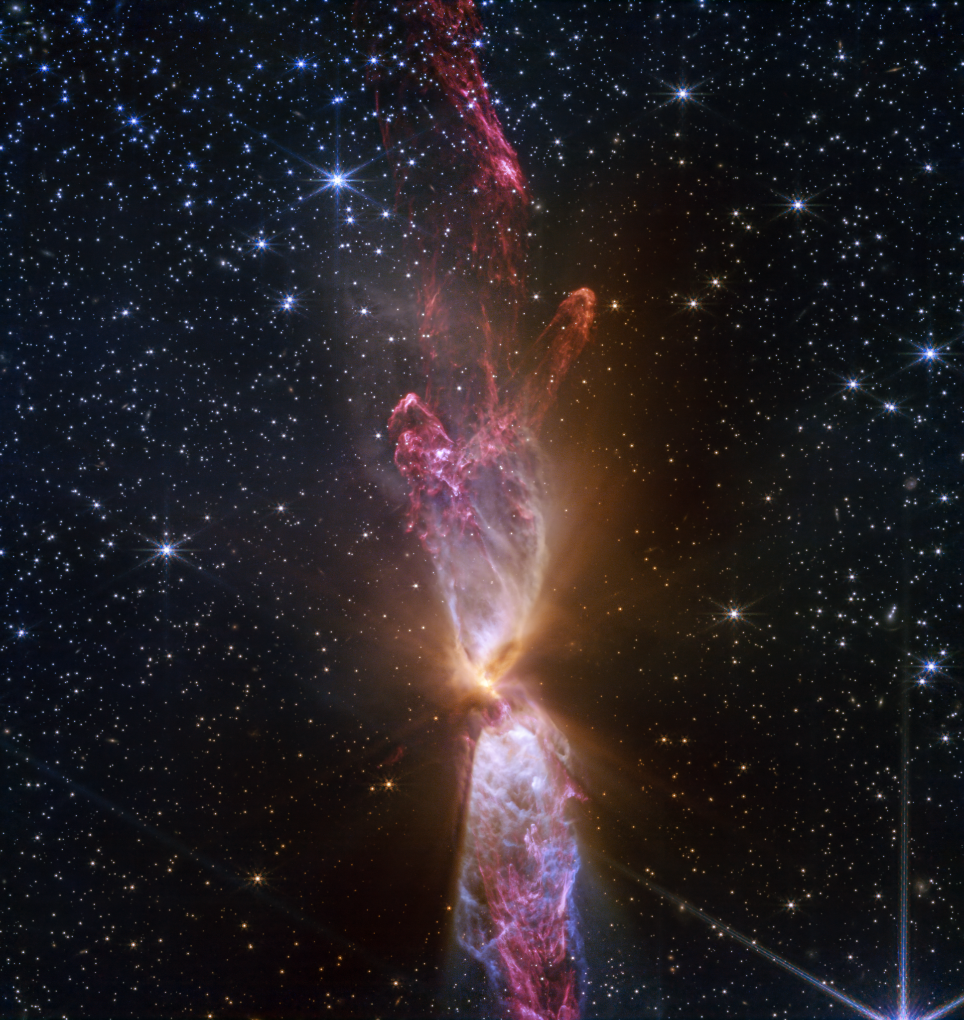 At the center is a thin vertical cloud known as Lynds 483 that is shaped like an hourglass with irregular edges. At lower center are two discrete bright white, tiny blobs of light that have raced away from the hidden central stars. The top lobe shows a more prominent orange U-shape. Orange bleeds into light purple, and brighter pink at its edges. Some background stars are visible through sections of this lobe. Higher up, there is an orange arc. Some brighter pink material extends to the top edges near the center. In the lower lobe, less orange is visible. More opaque light purple is in its top third, rippling out into semi-transparent blues and pinks. The lower lobe has more texture. V-shapes left and right of the lobes are darkest, and the background stars in these areas appear orange. Elsewhere, the black background of space is clearer, speckled with tiny white stars and faint orange galaxies.