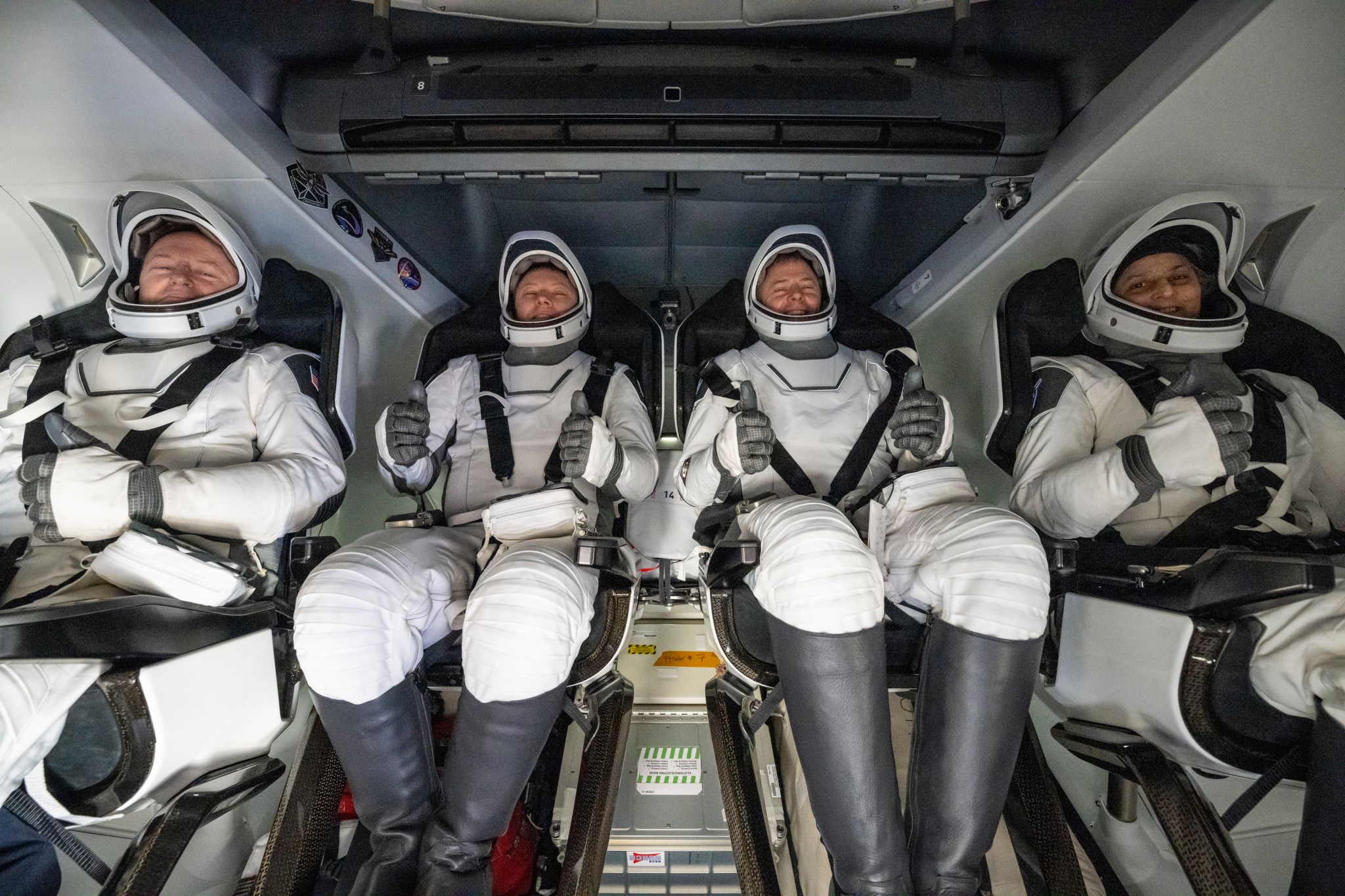 Four people in white spacesuits, white helmets, and black boots give a thumbs up from inside a SpaceX Dragon spacecraft.