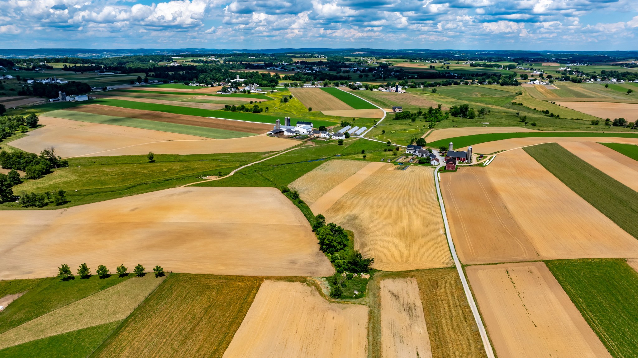 Aerial view of Amish countryside with patchwork fields in shades of tan, green, and brown. White farmhouses and barns dot the landscape under a blue sky with fluffy white clouds.