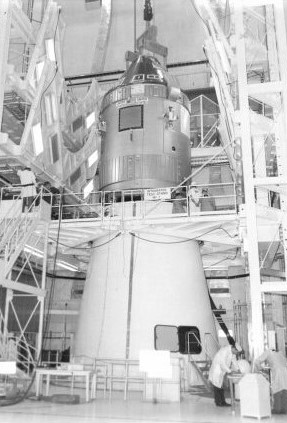 Black and white image of a silver spacecraft (Apollo) being lowerered onto a white truncated cone (Spacecraft LM Adapter) in a large assembly hall.