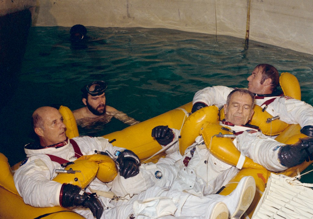 Image of three astronauts wearing white spacesuits sitting in yellow life rafts in what appears to be a circular indoor pool or water tank.