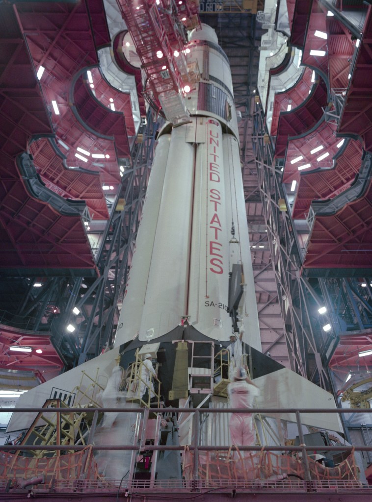 Image of the base of a large rocket taken from the bottom looking up, inside a large assembly building, with workers replacing stabilizing fins.