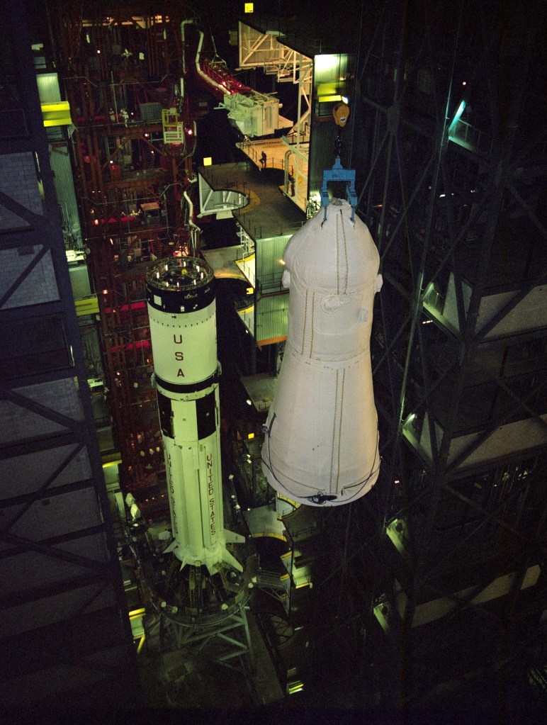 Workers prepare to lower a white spacecraft onto its rocket in a large assembly building.