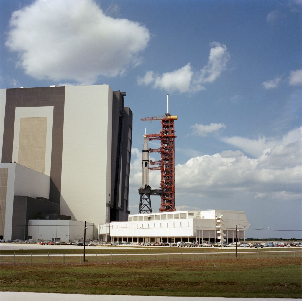 Distant view of a rocket standing vertically on its launch platform leaving a large assembly building.