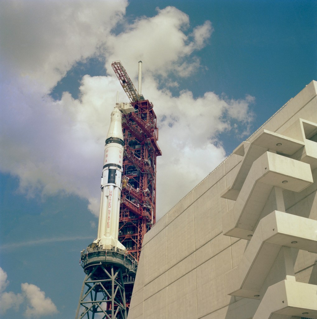 View from street level of a large white and black rocket and its red launch tower next to a four-story building.