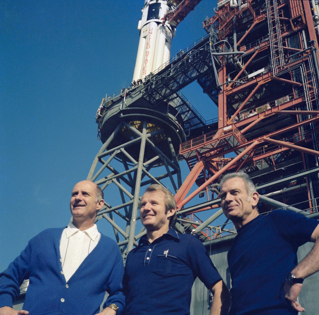 Image of three male astronauts wearing blue shirts standing in front of a large rocket, only its base is visible.