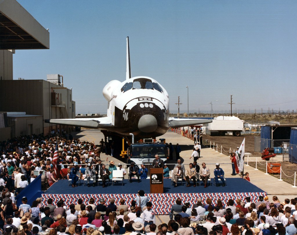 View of a space shuttle orbiter with a about a dozen people on a stage in front of it.