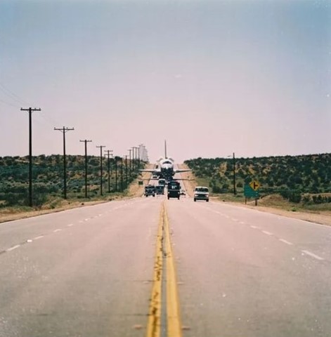 Distant view of a space shuttle orbiter being trucked on a desert highway.