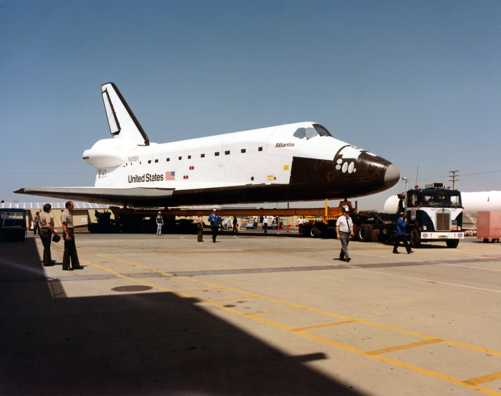 Image of a space shuttle orbiter on a runway apron being towed by a blue truck.