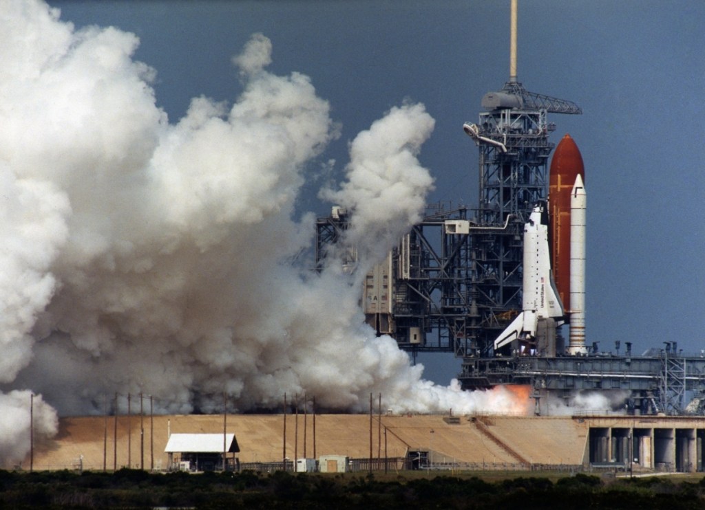 View of a space shuttle on its launch pad with its engines lighting up and smoke and steam billowing from the pad.