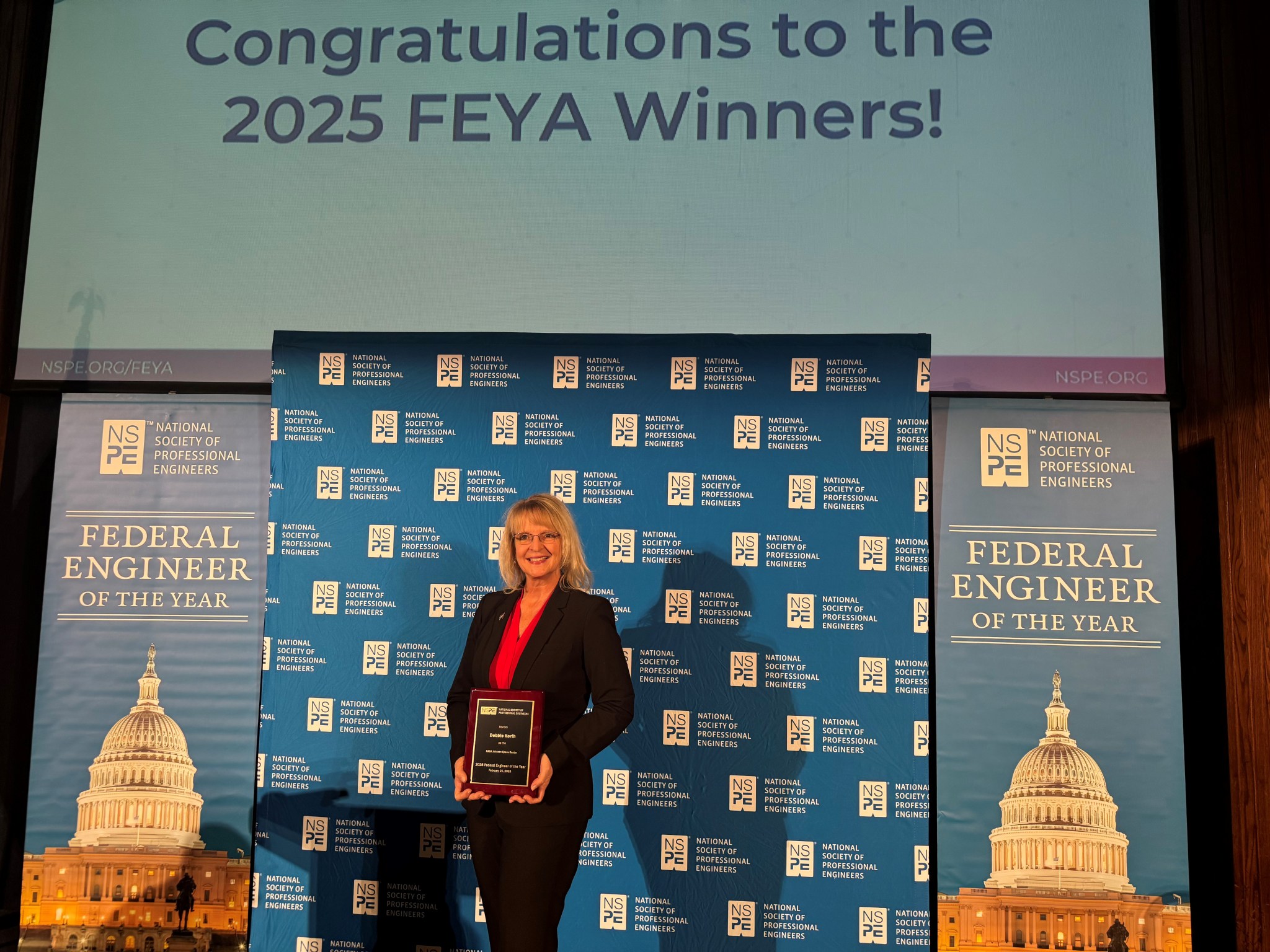 Orion Deputy Program Manager Debbie Korth, in a black suit and pink blouse, stands on a stage with the National Society of Professional Engineers logo behind her.