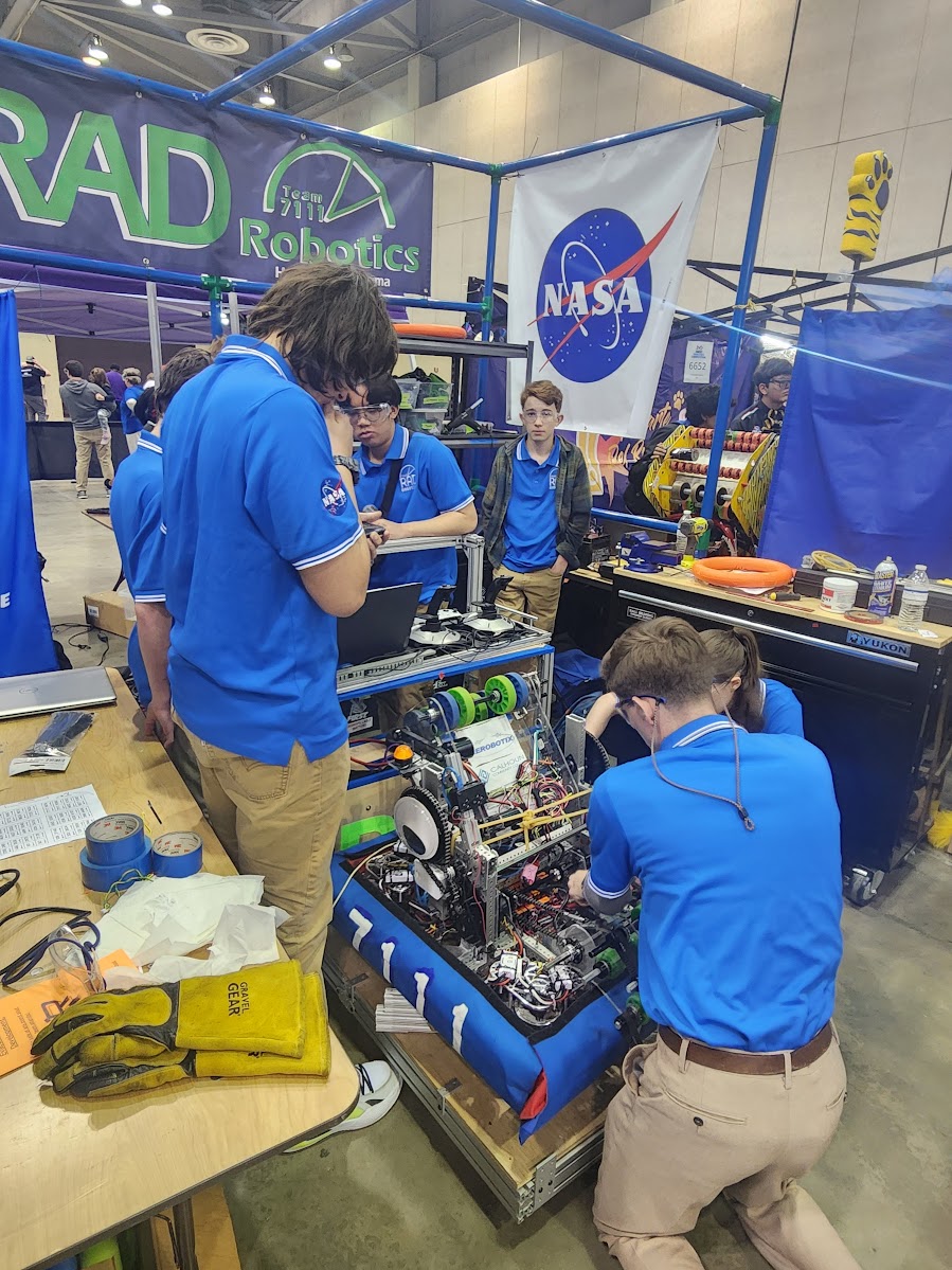 Students from RAD Robotics Team 7111 – a FIRST Robotics team from Huntsville, Alabama, stand in blue shirts working on their robot with a large NASA meatball logo in the background.