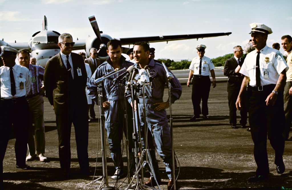 Two astronauts in blue flight suits speak at a microphone at an airport.