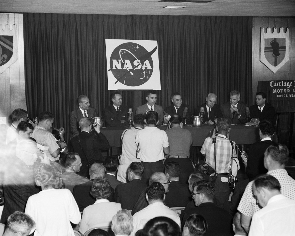 Black and white image of seven men sitting behind a long dais during a press conference.