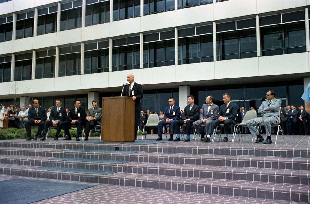 Image of a man in a suit standing at a podium with several others seated on either side in front of a building.