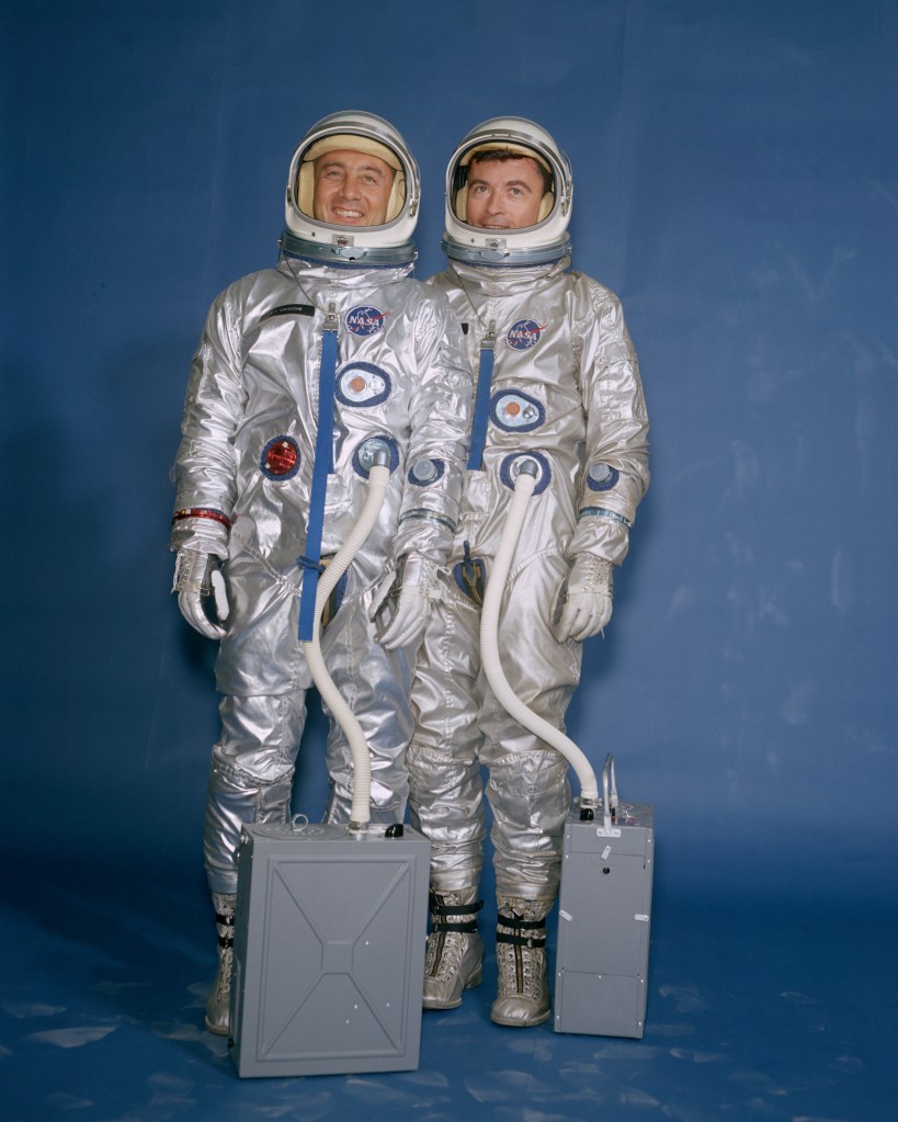 Image of two male astronauts wearing silver spacesuits and white helmets.