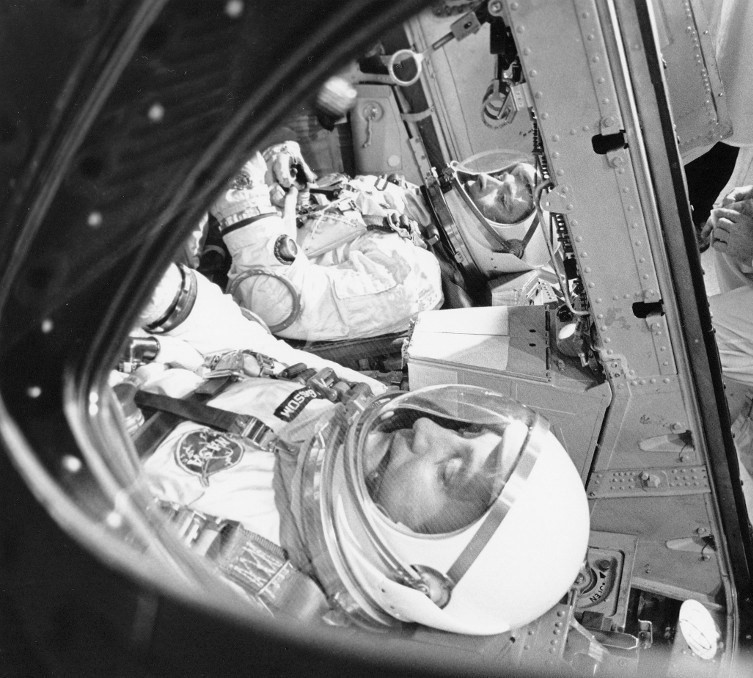 Black and white image of two spacesuited astronauts inside their capsule, seen through the capsule's window.