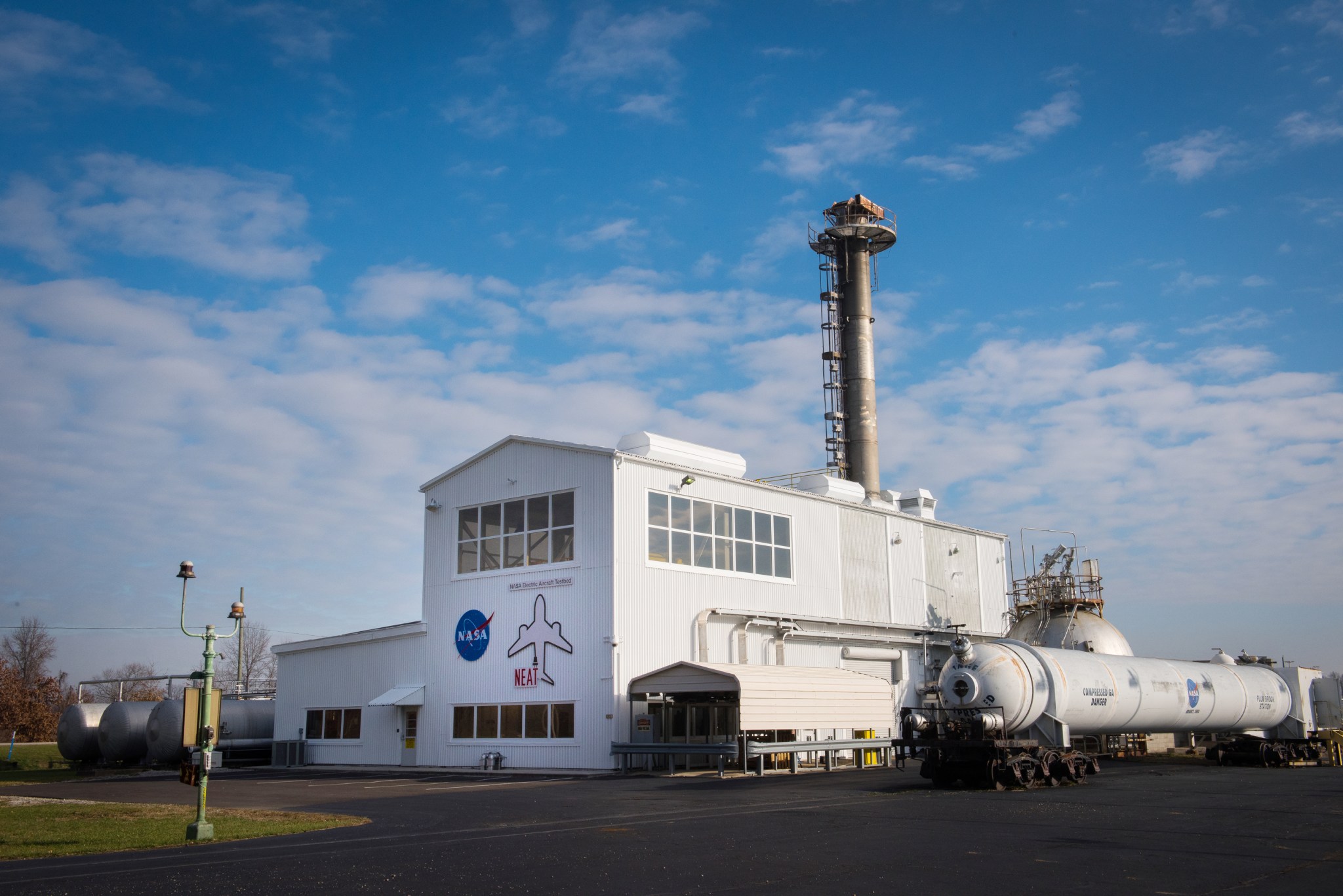 A metal building with an airplane and NASA logo on the front. A tall section is in the middle, a smaller flat section is on the left, and a large cylinder with tubing goes into the building on its right.