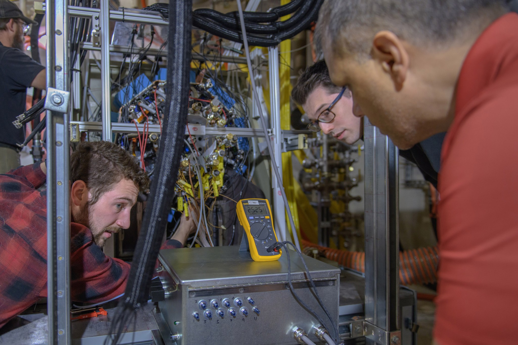 Three graduate researchers, one on the left and two on the right, look at a yellow unit monitoring a testbed of wiring and tubes.