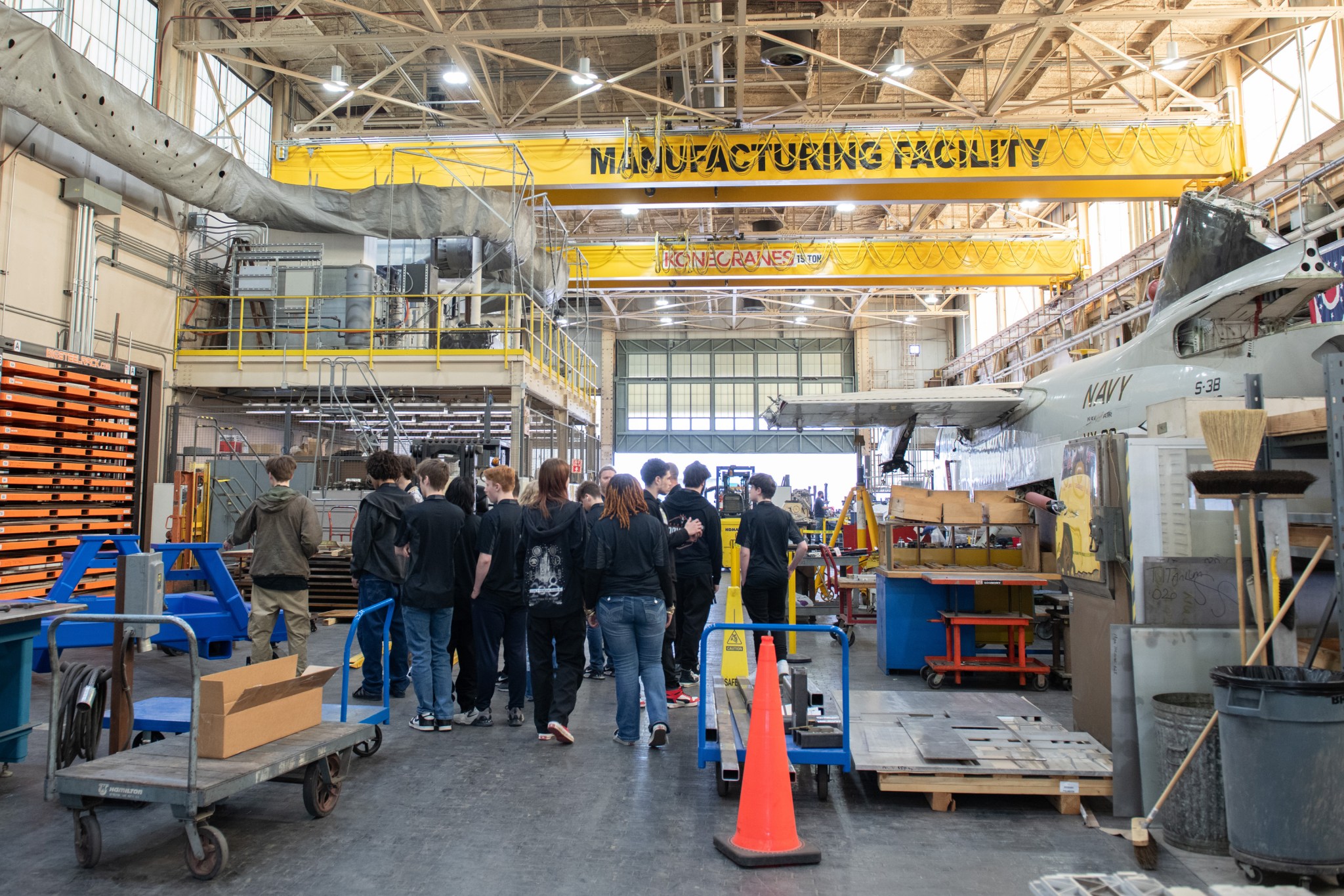 A group of students with their backs to the camera stand inside a large NASA facility. They are surrounded by equipment, including carts, boxes, safety cones, and other hardware. A large yellow beam above their heads shows text that reads, “MANUFACTURING FACILITY.”