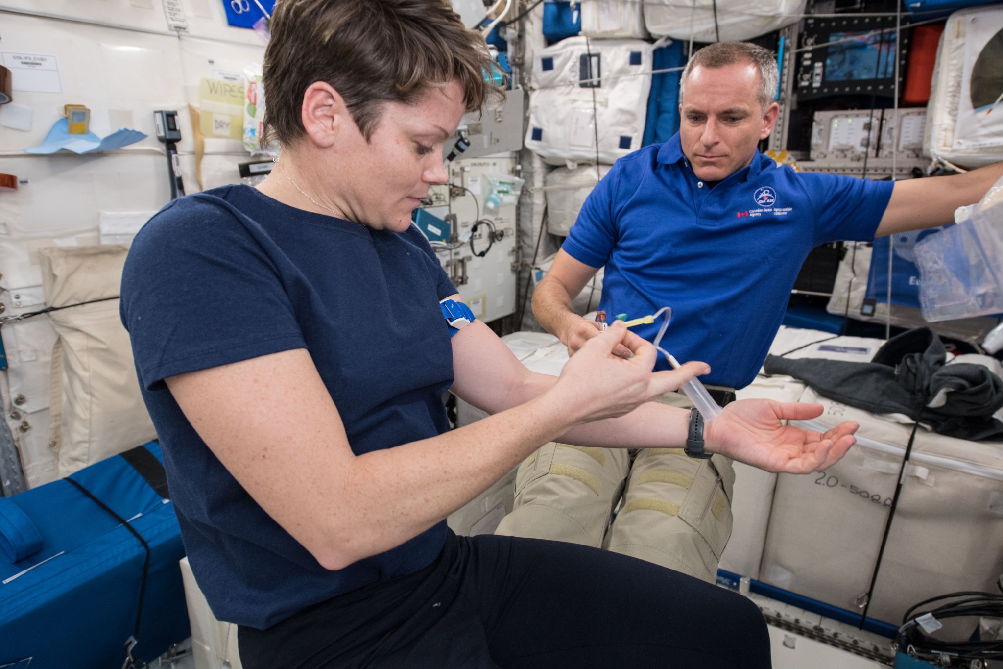 McClain, wearing a blue t-shirt and black pants, has a blue band around her upper left arm and holds a blood sample collection device in her right hand. Behind her is Saint-Jacques, wearing a blue polo shirt with a CSA logo and khaki pants and holding a sample vial in his right hand.