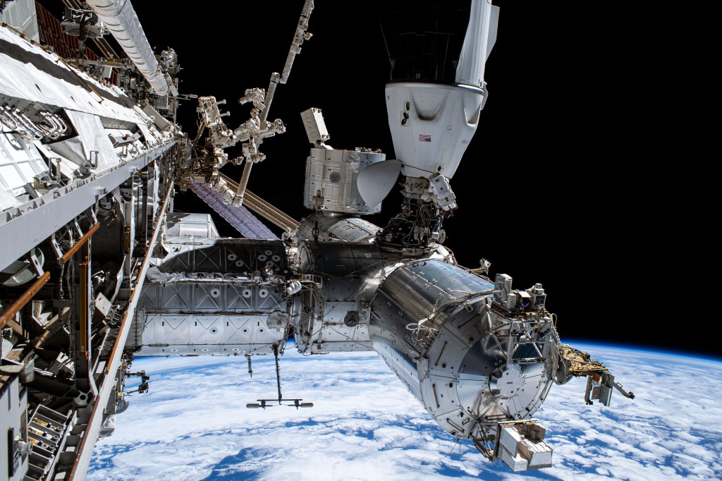 The view in this image shows the space station exterior from the vantage point of the starboard truss. the view shows the US segment, including the US Lab, Node 2, Columbus, and JEm modules. The SpaceX Dragon Crew-9 vehicle is docked to the Node 2 zenith port. The Earth limb is in the background. A crew member in a spacesuit can be seen outside the Dragon vehicle.