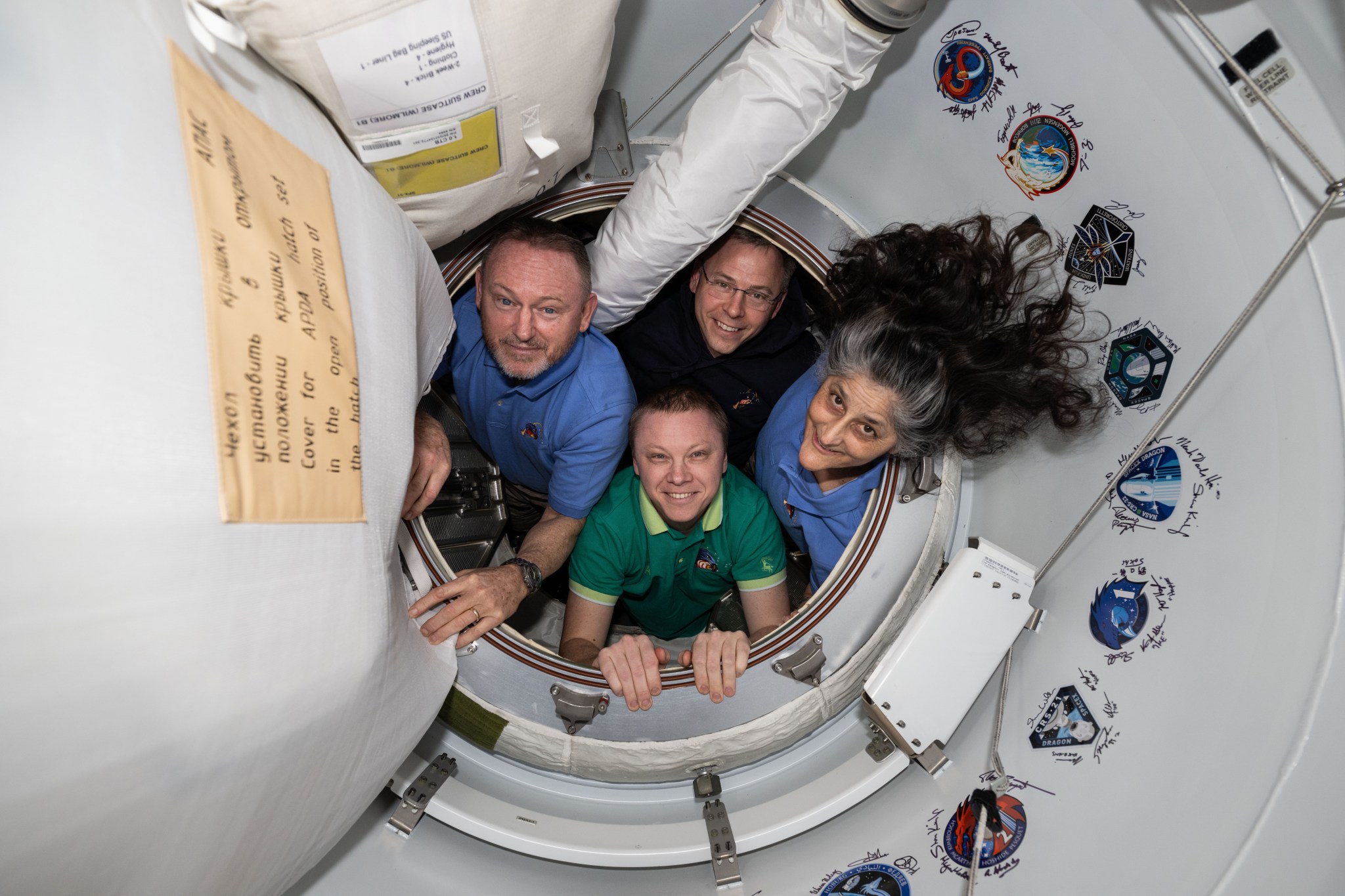 Four people are in a circular opening, smiling at the camera. They are wearing polo shirts in different colors. Suni Williams' (far right) hair spills outward toward a display of patches.