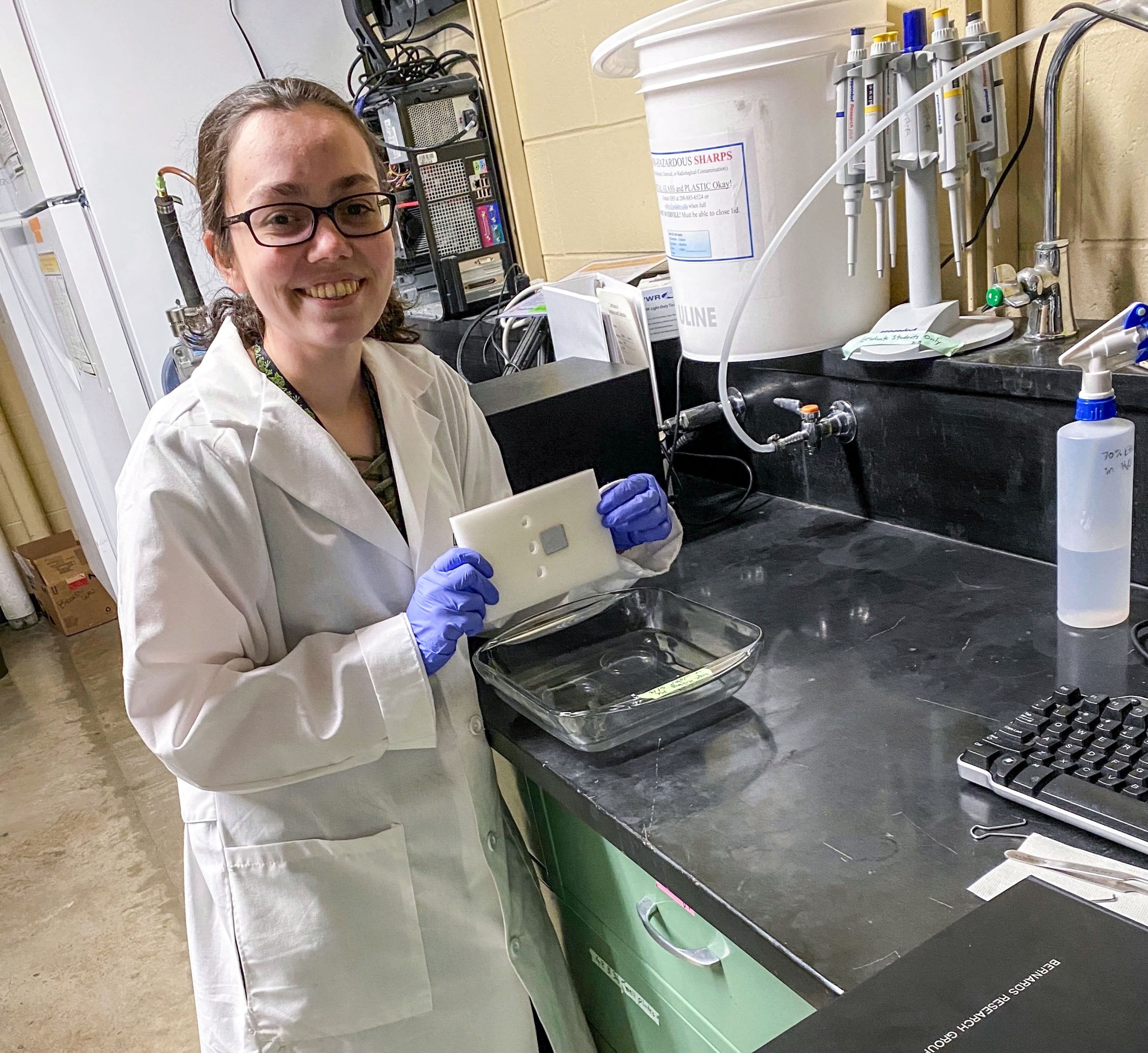 A woman stands in a white lab coat and smiles while holding a white rectangular sample in a laboratory.