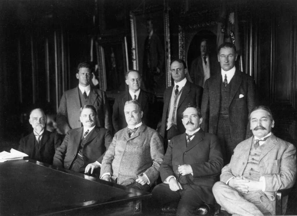 Black and white photograph of a group of nine men in suits, five seated, four standing.