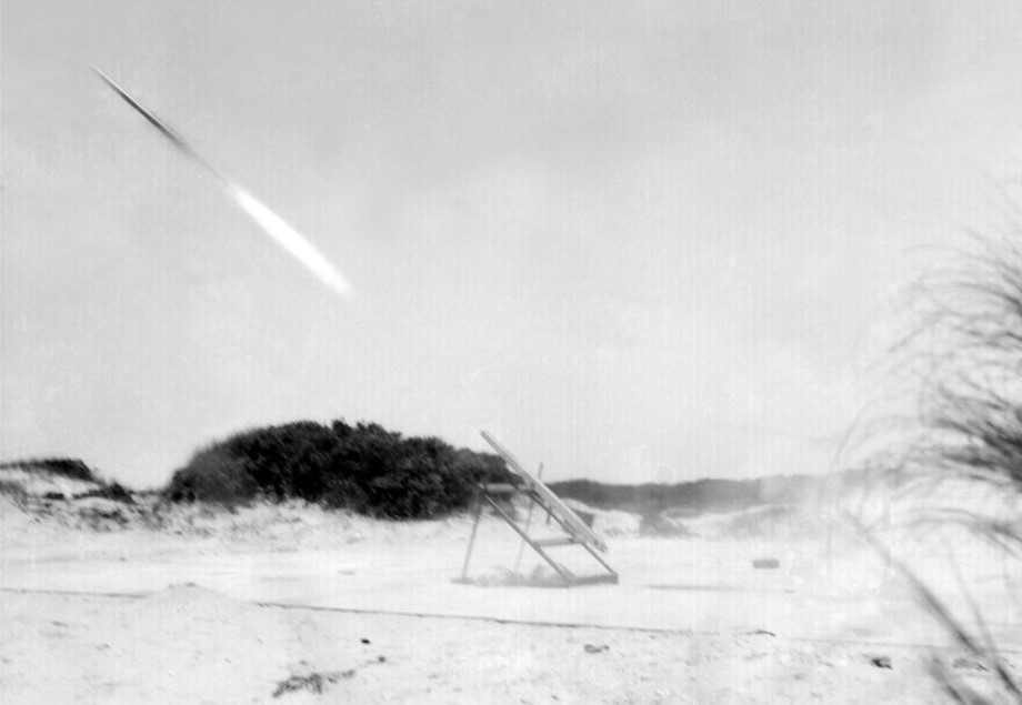 Black and white photograph of a sounding rocket taking off from a beach.