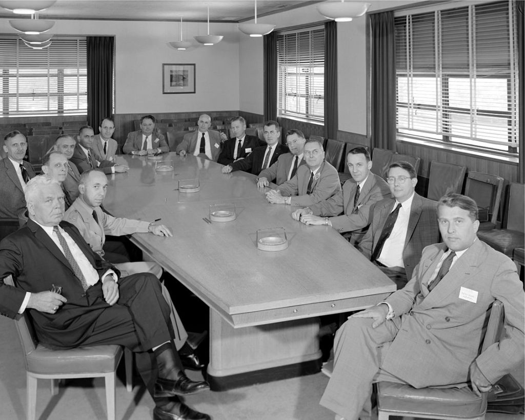 Black and white group photograph of about 20 men in suits sitting around a large conference table.