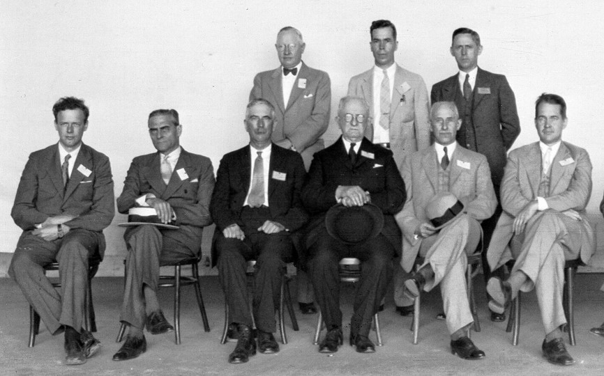 Black and white photograph of nine men wearing suits, six seated in front, three standing behind them.