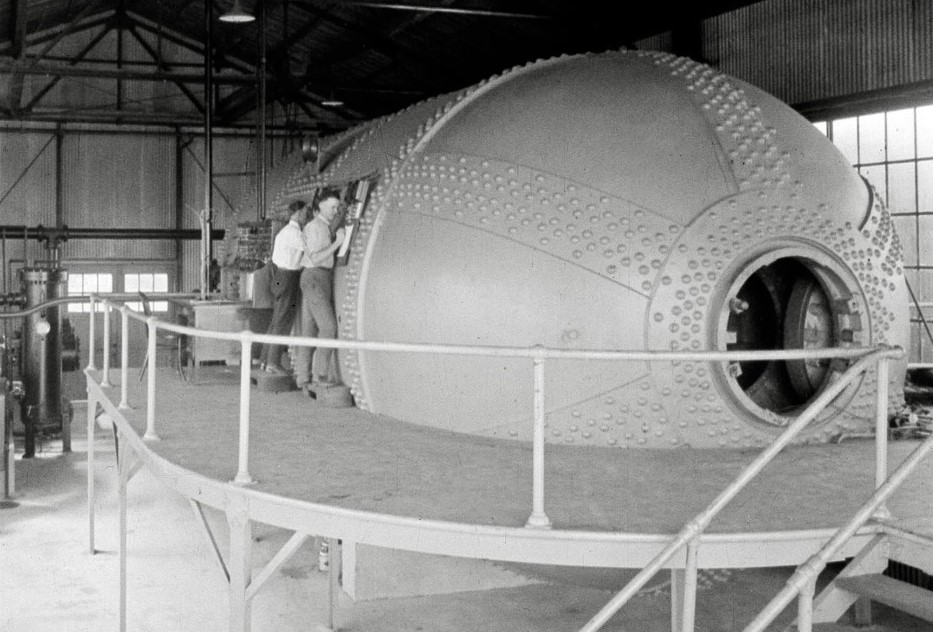 Black and white image of two men peering into the side of a large cylindrical object (a wind tunnel).