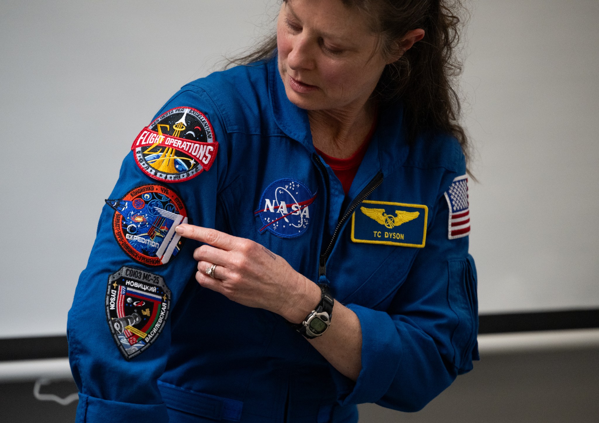 Astronaut Tracy Dyson points to the Expedition 71 patch on her right sleeve. The round patch has an orange border with a large, stylized "71" on the right side. She is wearing a blue flight suit with multiple patches.