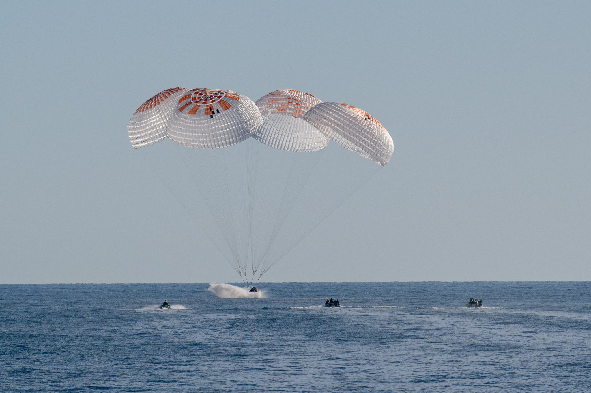 Four large white and red parachutes are fully deployed above a SpaceX Dragon spacecraft splashing into the water.