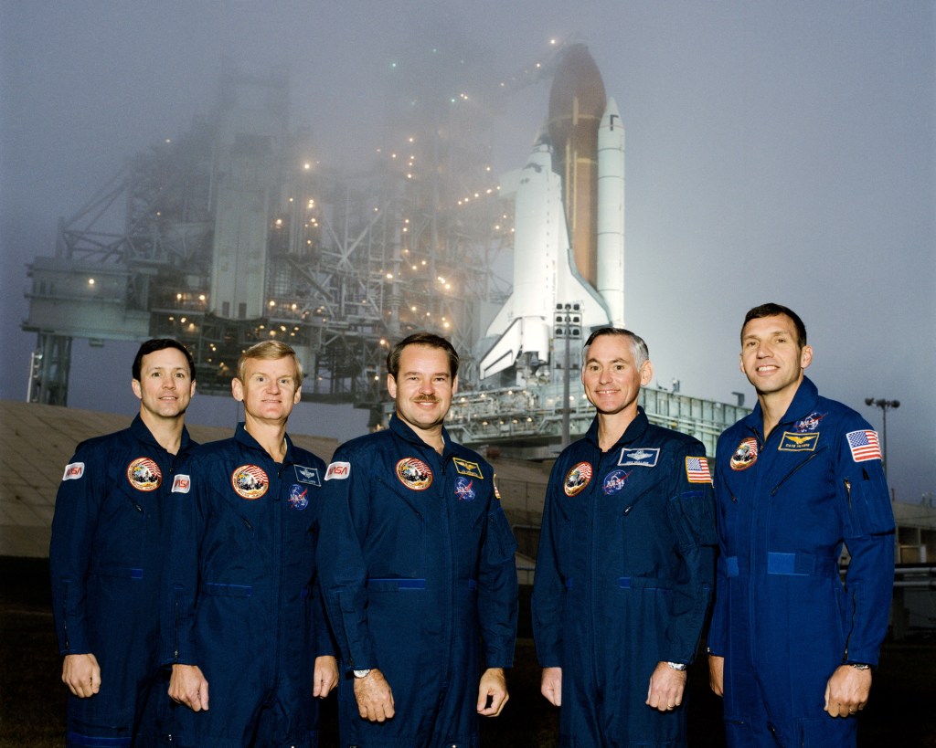 Group photograph of five astronauts wearing blue suits posed in front of a space shuttle on the pad.