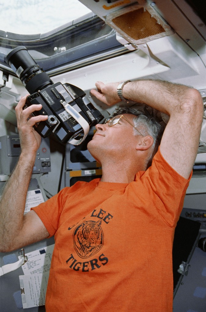 Photograph of an astronaut wearing an orange T-shirt taking a picture through a shuttle window.