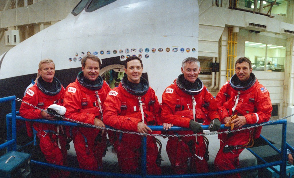 A group photo of five astronauts wearing orange spacesuits without helmets, standing in front of space shuttle trainer.