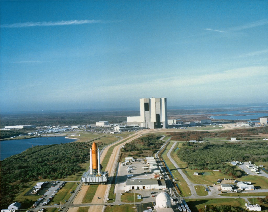 Aerial view of a space shuttle being transported on a crawler from the assembly building visible in the background to the launch pad.