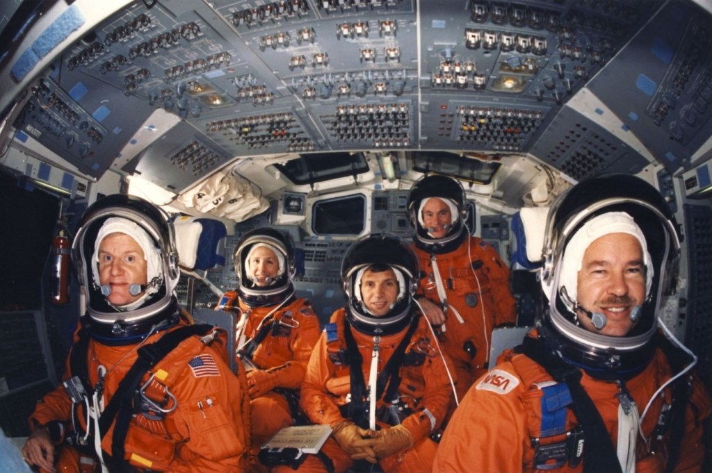 Group photo of five astronauts wearing orange space suits with helmets in a space shuttle cockpit simulator