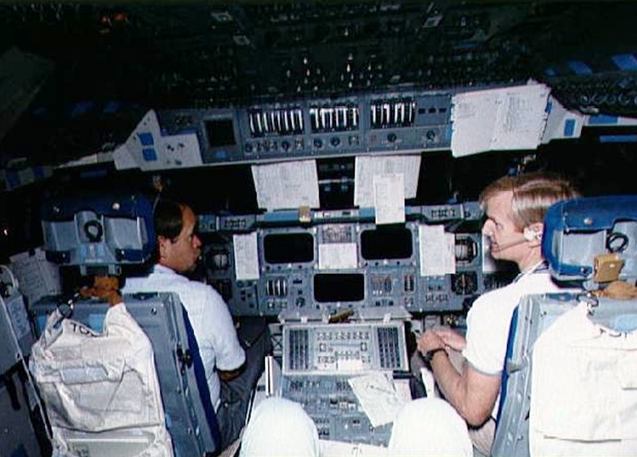 View of inside of shuttle cockpit simulator with two astronauts in the commander's and pilot's seats.