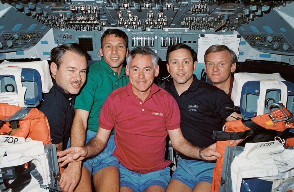 A group photo of five astronauts wearing different color polo shirts on the flight deck of a space shuttle.