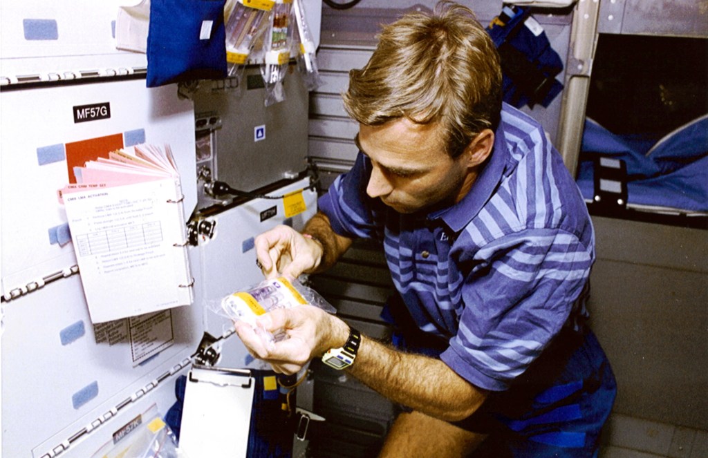 Image of a male astronaut in a blue and white polo shirt working on an experiment in the shuttle middeck.