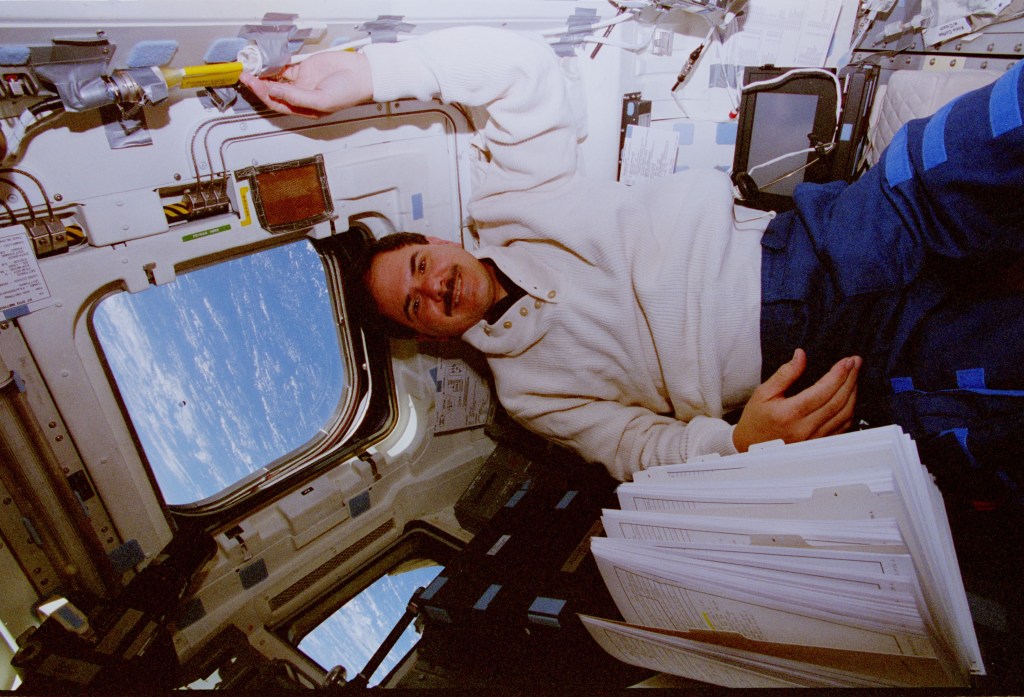 Image of a male astronaut floating near a shuttle overhead window.