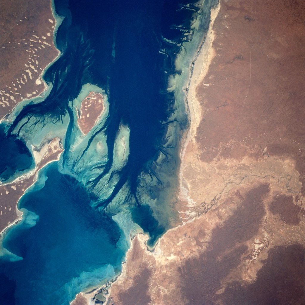 View from space of a desert coastline on the right, with a blue water channel with several islands on the left.