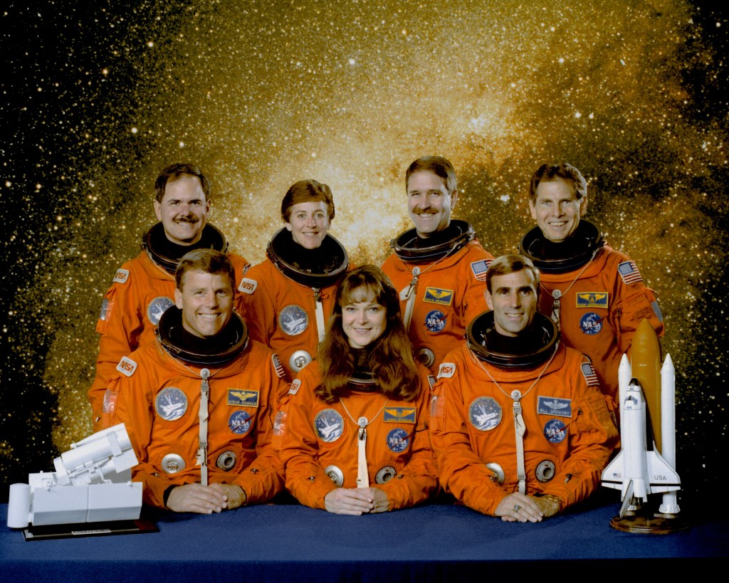 Group photo of seven astronauts wearing orange spacesuits with no helmets, with a galaxy in the background.